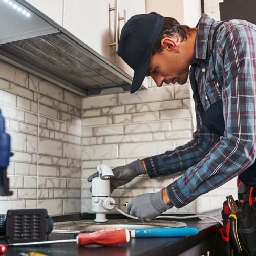 Electric faucet. Rear view of handsome plumber repairing sink in kitchen with tools box