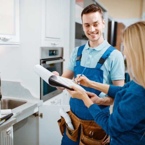 Male plumber in uniform and female customer in the kitchen. Handyman with toolbag repair sink, sanitary equipment service at home